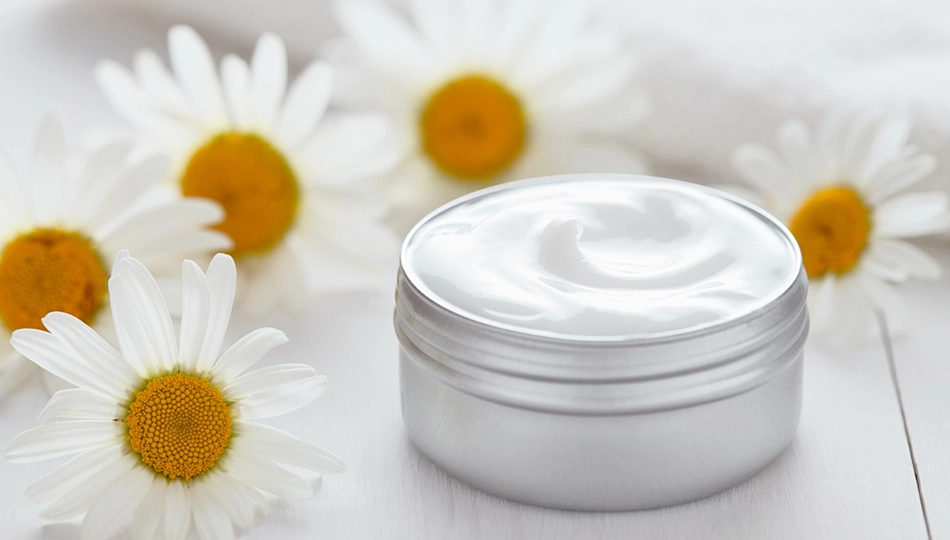 Naturkosmetik Unreine Haut Tiegel mit Gesichtscreme auf weißem Holzuntergrund, umgeben von Kamillenblüten.