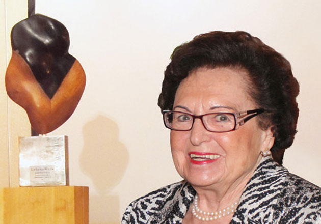 Ältere Frau mit Brille und Perlenkette lächelt neben einer abstrakten Holzskulptur auf einem Sockel