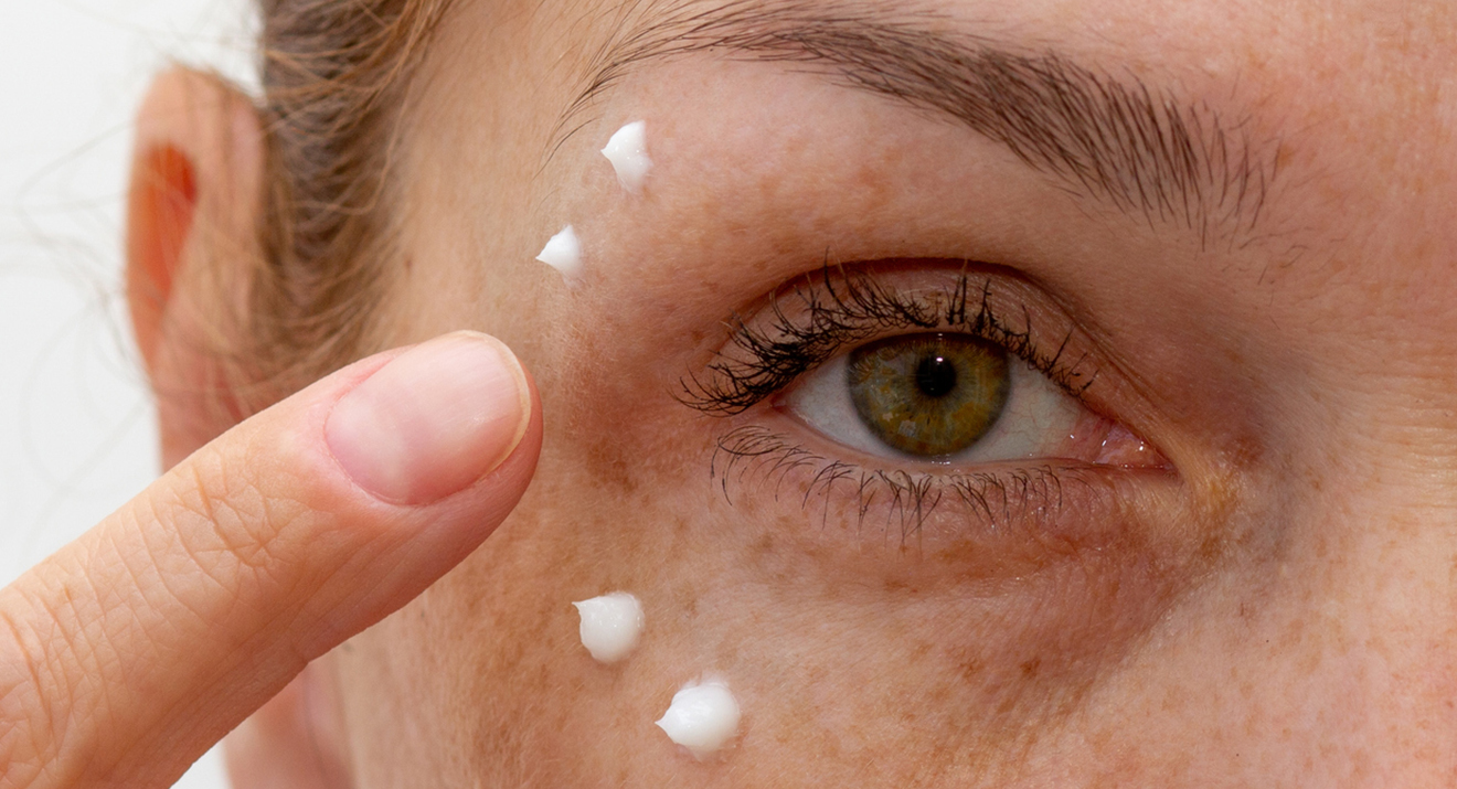 Augencreme für empfindliche Augen: Nahaufnahme eines offenen grünen Auges, umgeben von aufgetupften Punkten Augencreme, die sanft mit dem Finger aufgetragen wird.
