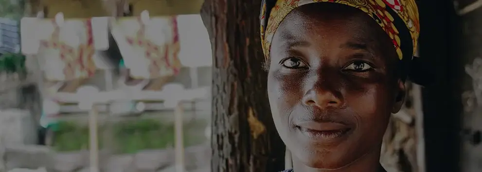 Frau mit buntem Kopftuch steht vor Baum, im Hintergrund unscharfe Wäscheleine mit bunten Tüchern