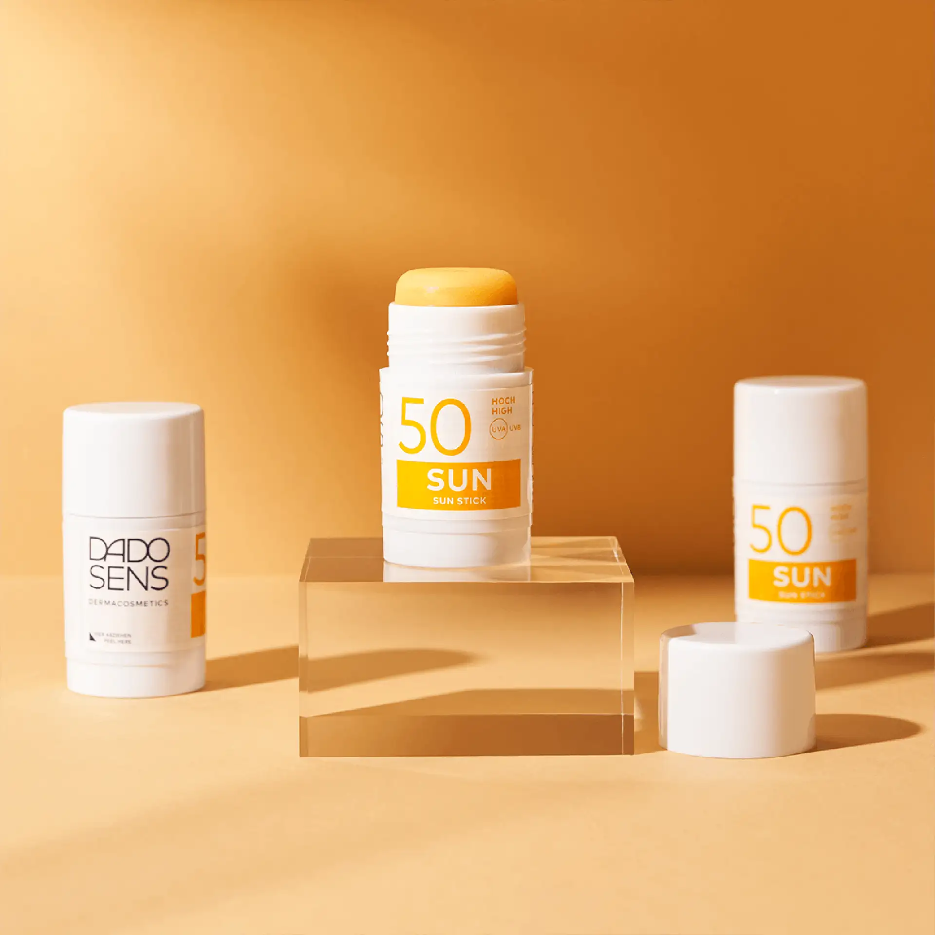 DADO SENS Sun Stick SPF 50 mit offenem Stift auf transparentem Podest, zwei geschlossene Sticks im Hintergrund