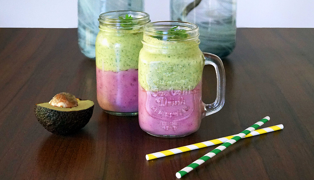 Zwei Gläser mit grünem und lila Avocado-Smoothie, halbe Avocado und bunte Trinkhalme auf Holztisch