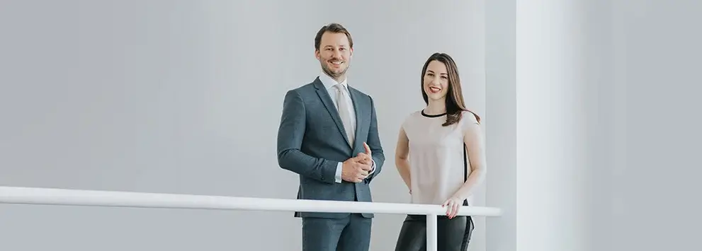 Zwei Personen in Businesskleidung stehen an einem weißen Geländer vor hellem Hintergrund und lächeln in die Kamera