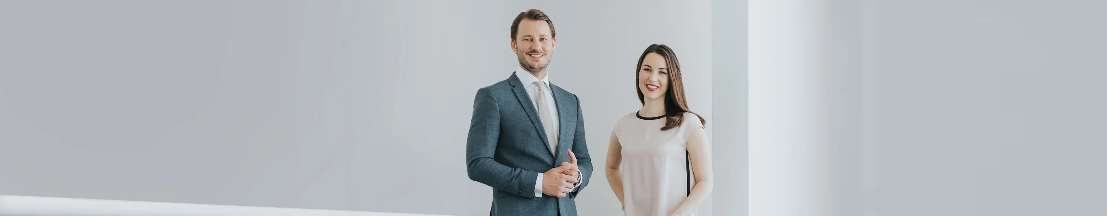 Zwei Personen in Businesskleidung stehen lächelnd in einem hellen, modernen Büro mit weißen Wänden