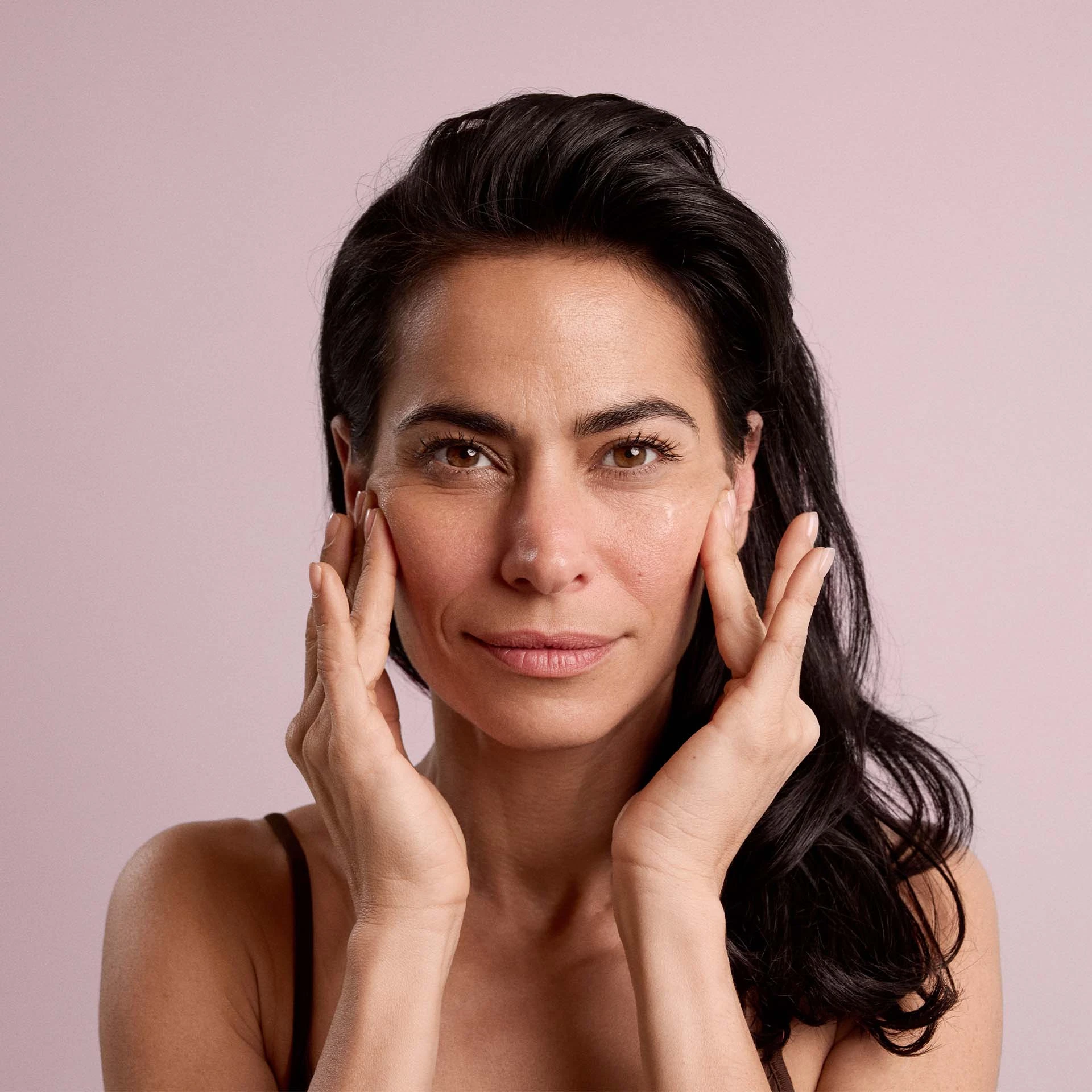 Frau berührt mit beiden Händen ihr Gesicht, neutraler Gesichtsausdruck vor rosa Hintergrund