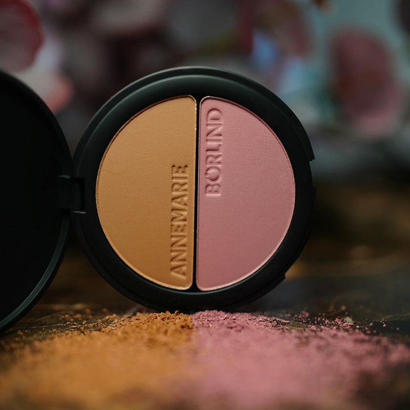 ANNEMARIE BÖRLIND 2in1 Bronzer & Blush mit warmen Herbsttönen aus der Make-up Kollektion „A Hug of Colour“ auf dunkler Oberfläche mit verstreutem Puder