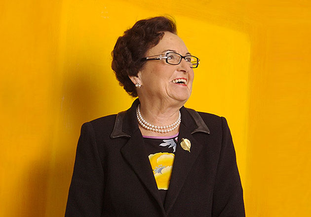 Annemarie Lindner mit Brille und Perlenkette lächelt seitlich vor gelbem Hintergrund