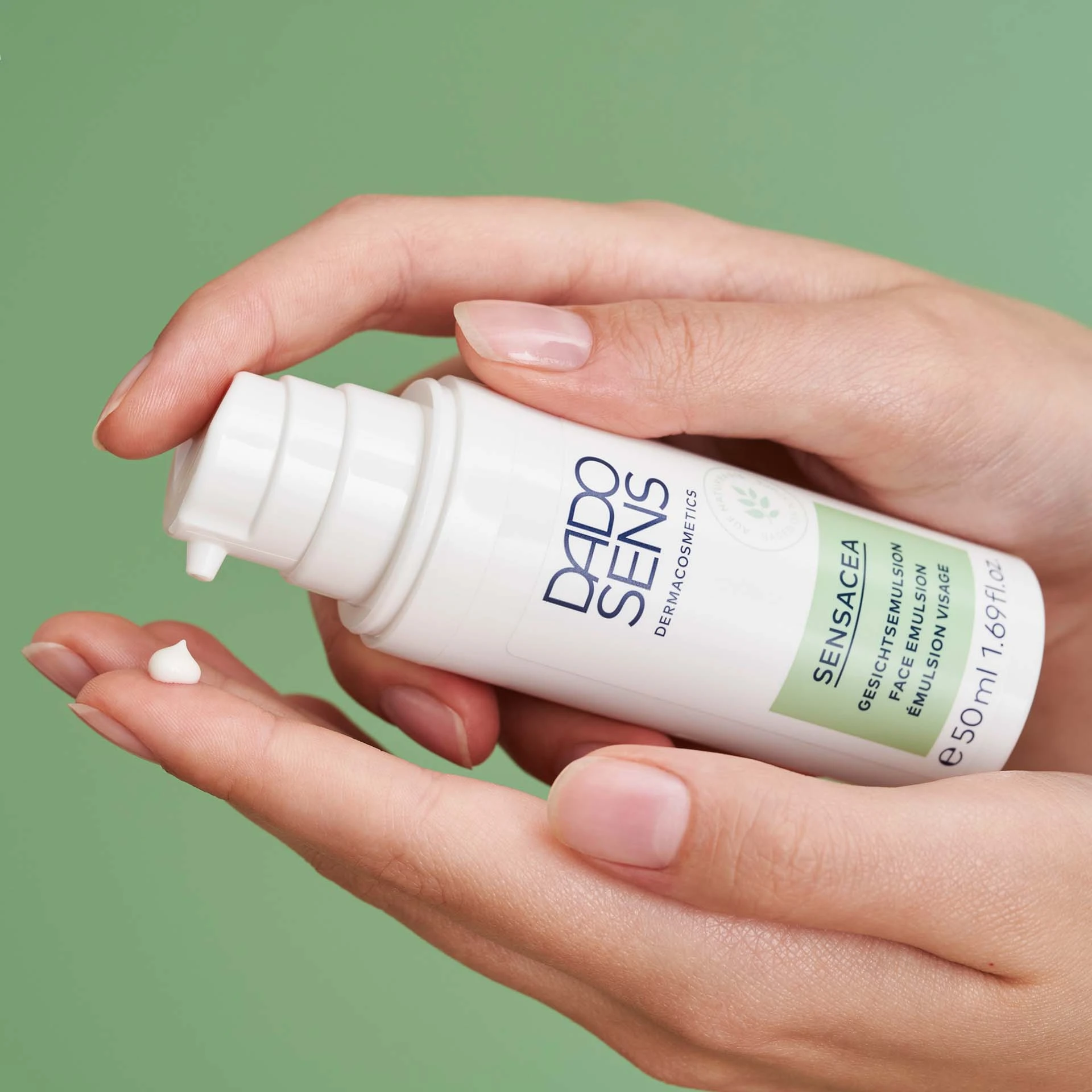 DADO SENS SENSACEA Gesichtsemulsion in weißer Flasche, weiße Creme auf Finger vor grünem Hintergrund