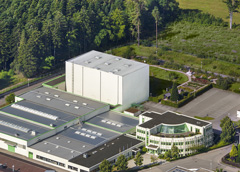 Luftaufnahme eines Industriegebäudes mit Solaranlagen auf dem Dach, umgeben von Wald und Parkplätzen