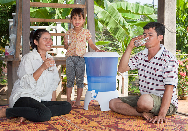 Familie sitzt auf Teppich im Freien, trinkt Wasser aus Gläsern neben blauem Wasserfilterbehälter