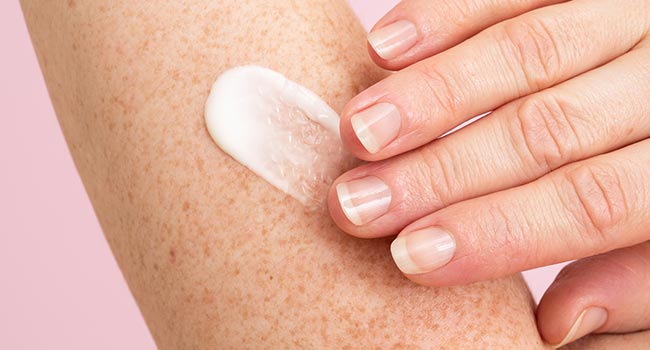 Hand cremt mit weißer Lotion einen Unterarm mit Sommersprossen ein vor rosa Hintergrund
