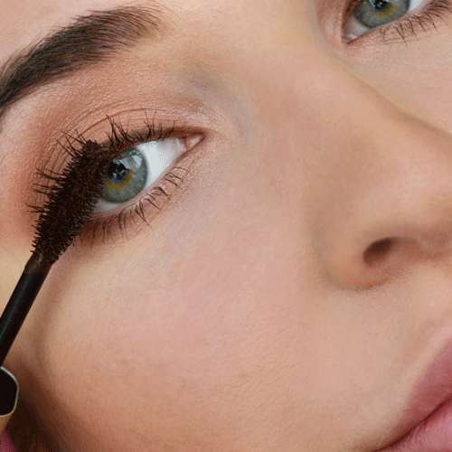 Nahaufnahme eines Auges mit grün-blauem Iris, Mascara wird auf die oberen Wimpern aufgetragen
