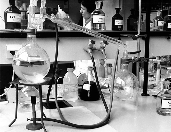 Labor mit Glasgeräten, Kolben, Schläuchen und Chemikalienflaschen, Person im Hintergrund bei Experiment