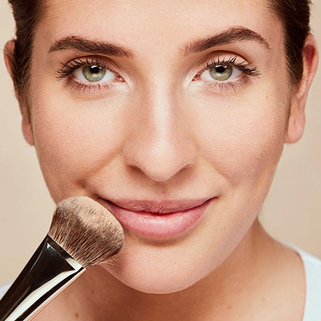 Frau trägt mit Pinsel Make-up auf Wange auf, Blick direkt in die Kamera, neutraler Hintergrund