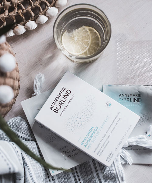 ANNEMARIE BÖRLIND Hyaluron Augenpads mit Sofort-Effekt neben Glas mit Zitronenscheiben und Textilien