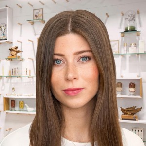 Nahaufnahme einer jungen Frau mit langen braunen Haaren und natürlichem Make-up vor weißem Regal mit Deko und Produkten