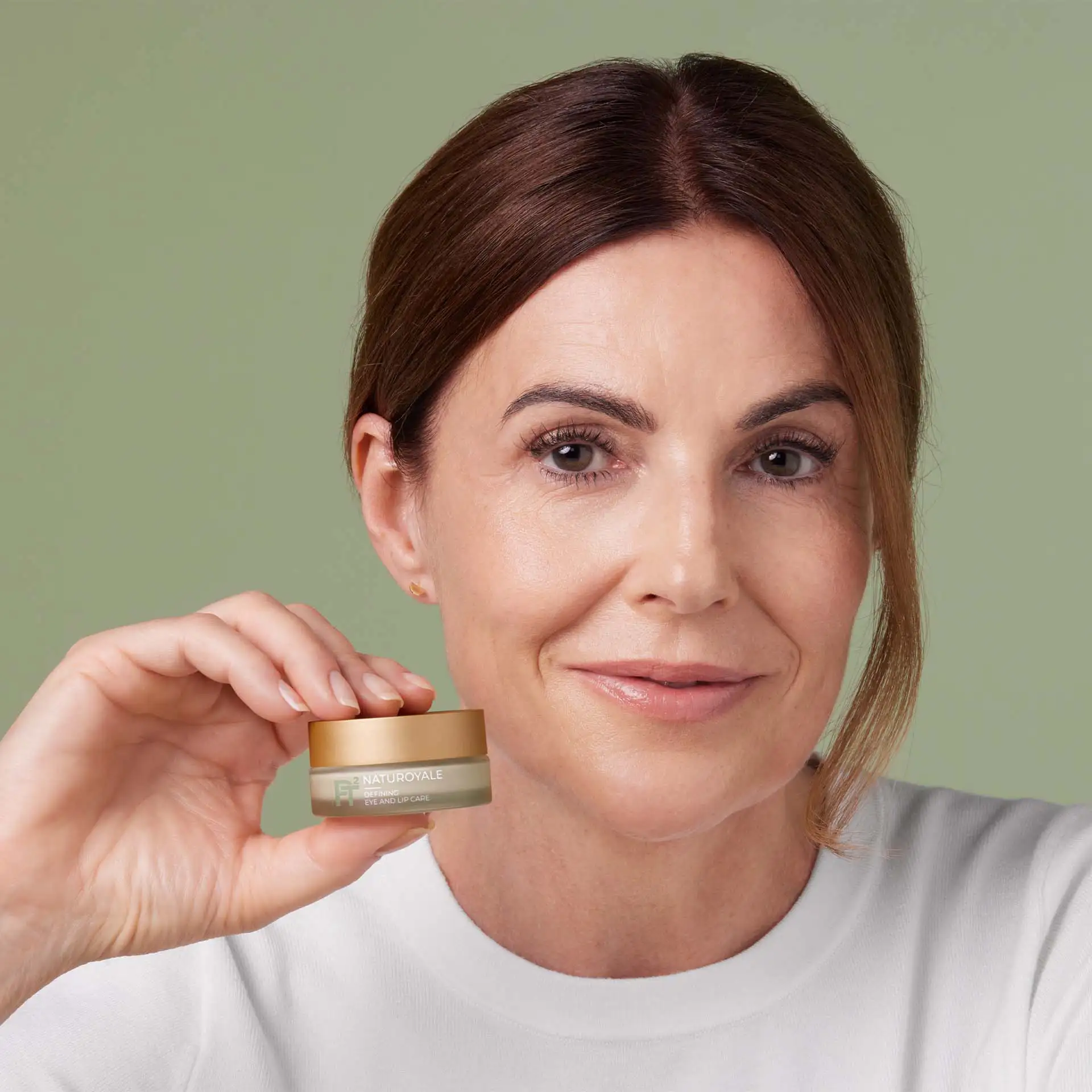 Frau hält Tiegel mit ANNEMARIE BÖRLIND NATUROYALE Defining Eye and Lip Care vor grünem Hintergrund