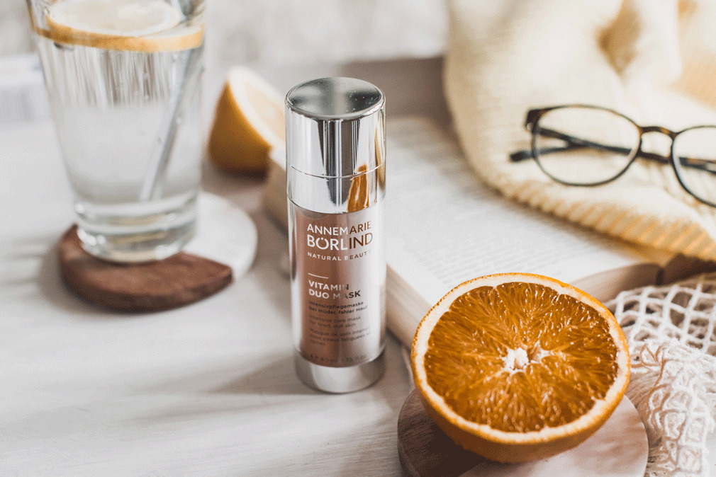 ANNEMARIE BÖRLIND Vitamin Duo Mask mit Orangenhälfte, Glas Wasser, Buch, Brille und Strickdecke
