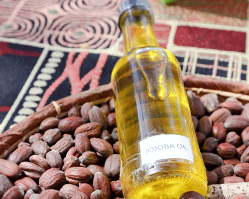 Flasche mit Jojobaöl liegt auf einem Korb voller Jojobasamen vor buntem Stoffmuster