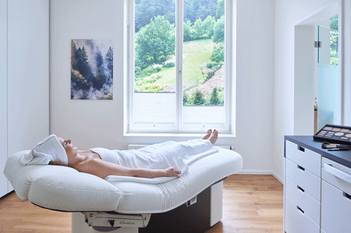 Frau entspannt auf Behandlungsliege in hellem Spa-Raum mit Blick auf grüne Landschaft durch Fenster