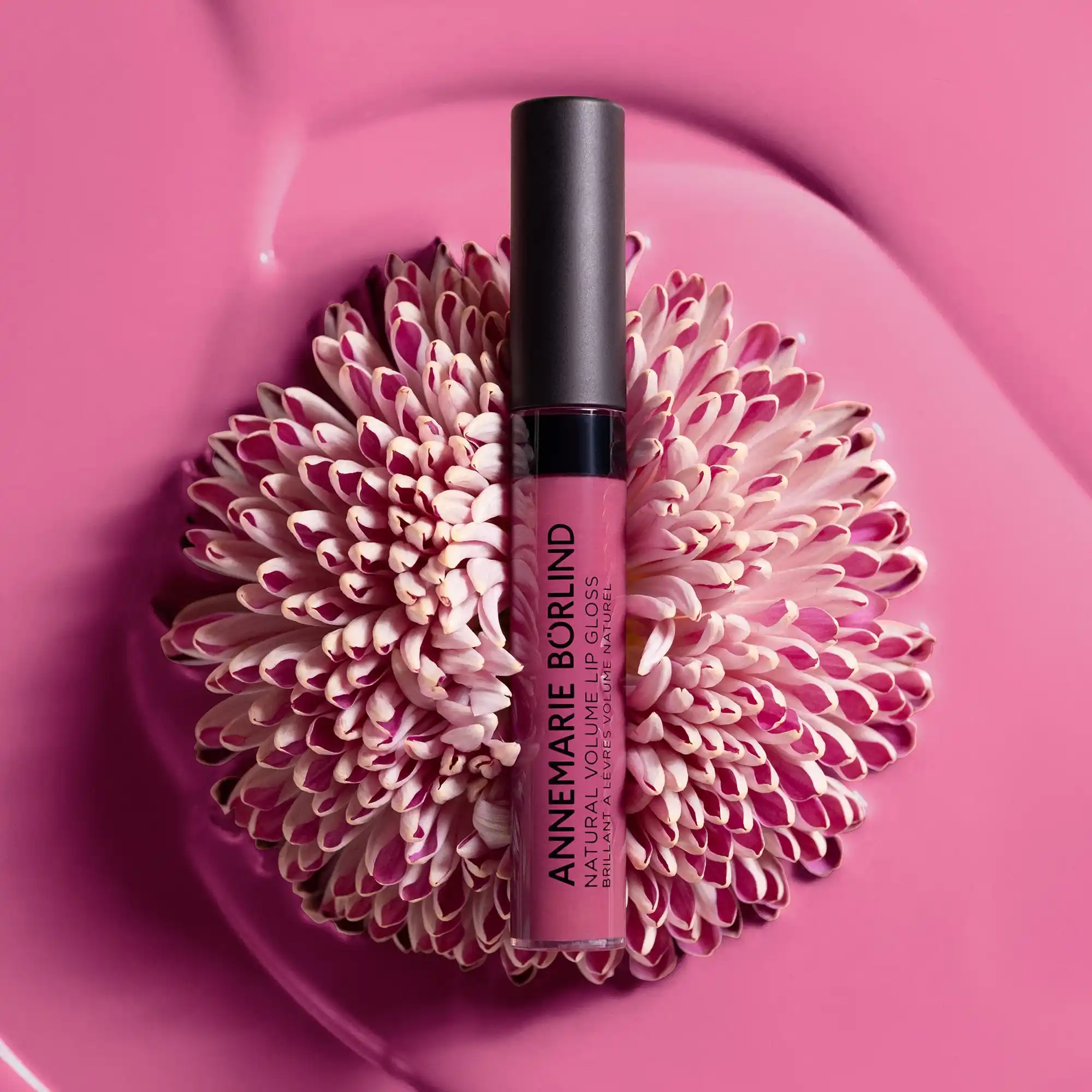 ANNEMARIE BÖRLIND NATURAL VOLUME LIP GLOSS in Farbe Snuggle Berry, dekorativ auf Blüte mit rosa und weißen Blütenblättern vor pinkem Hintergrund inszeniert