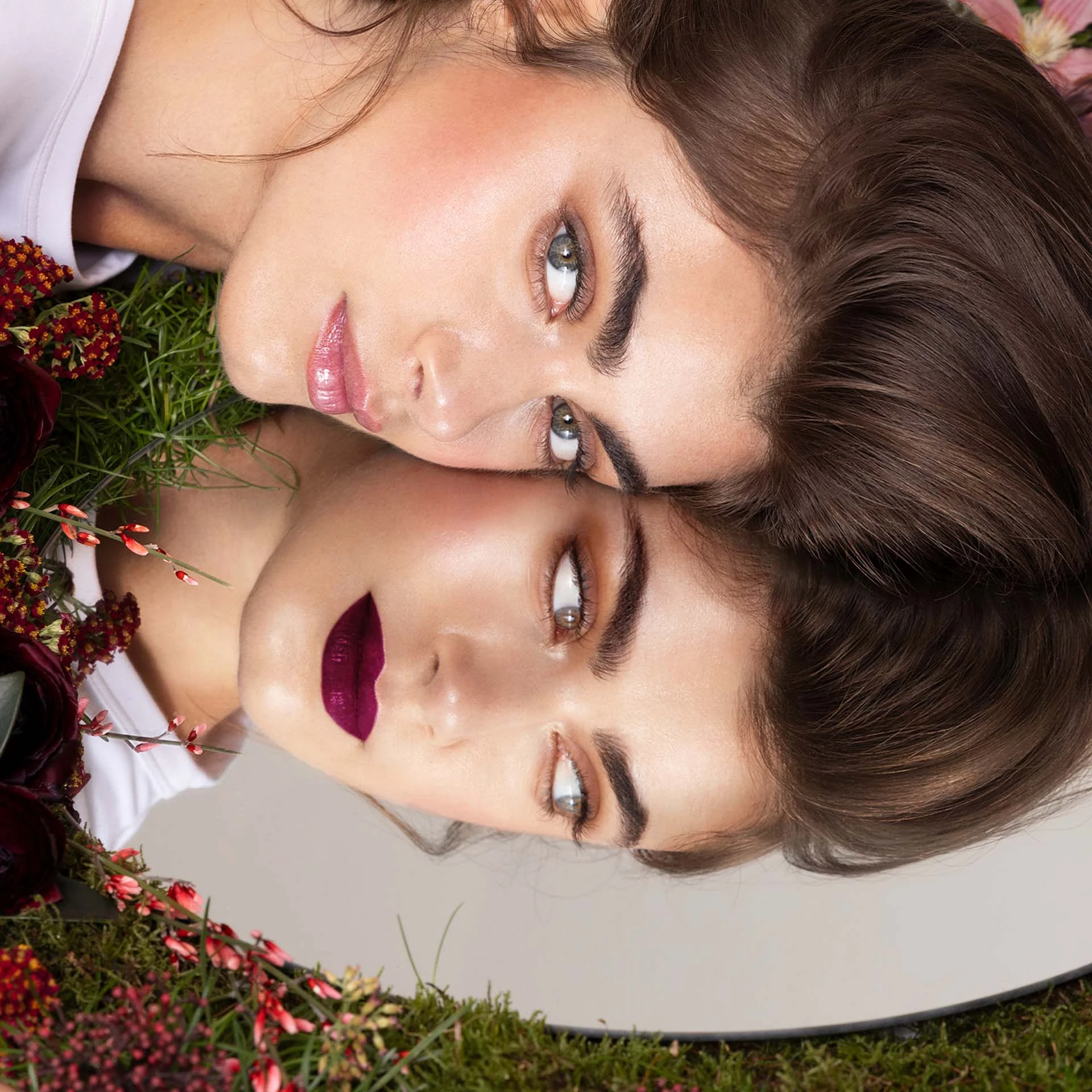 Frau mit natürlichem Make-up und Spiegelbild mit dunklem Lippenstift, umgeben von Blumen und Moos