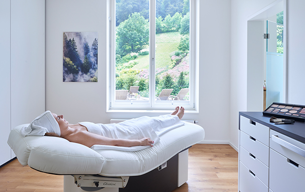 Frau liegt entspannt auf Behandlungsliege in hellem Raum mit Fenster und Naturblick im Spa