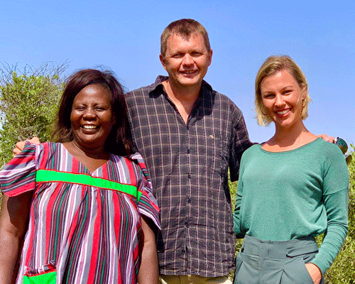 Drei Personen stehen lächelnd im Freien vor grünen Büschen und blauem Himmel, entspannt und freundlich