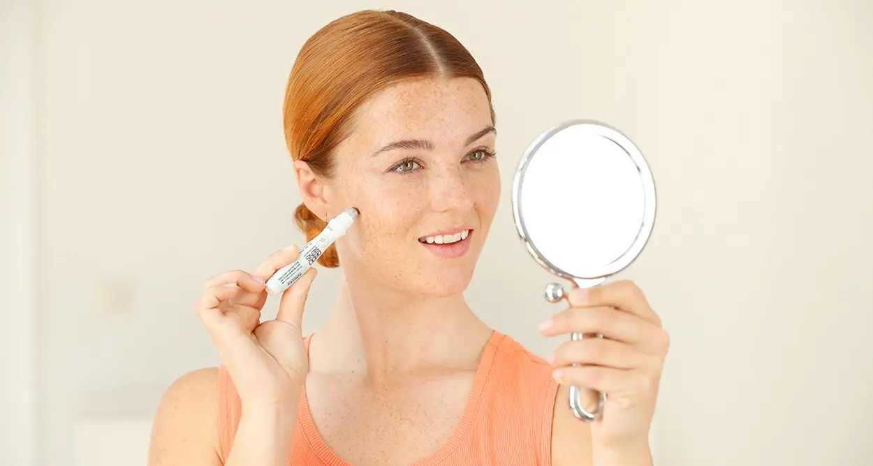 Frau trägt mit Applikator ein Hautpflegeprodukt auf die Wange auf und schaut in einen Handspiegel