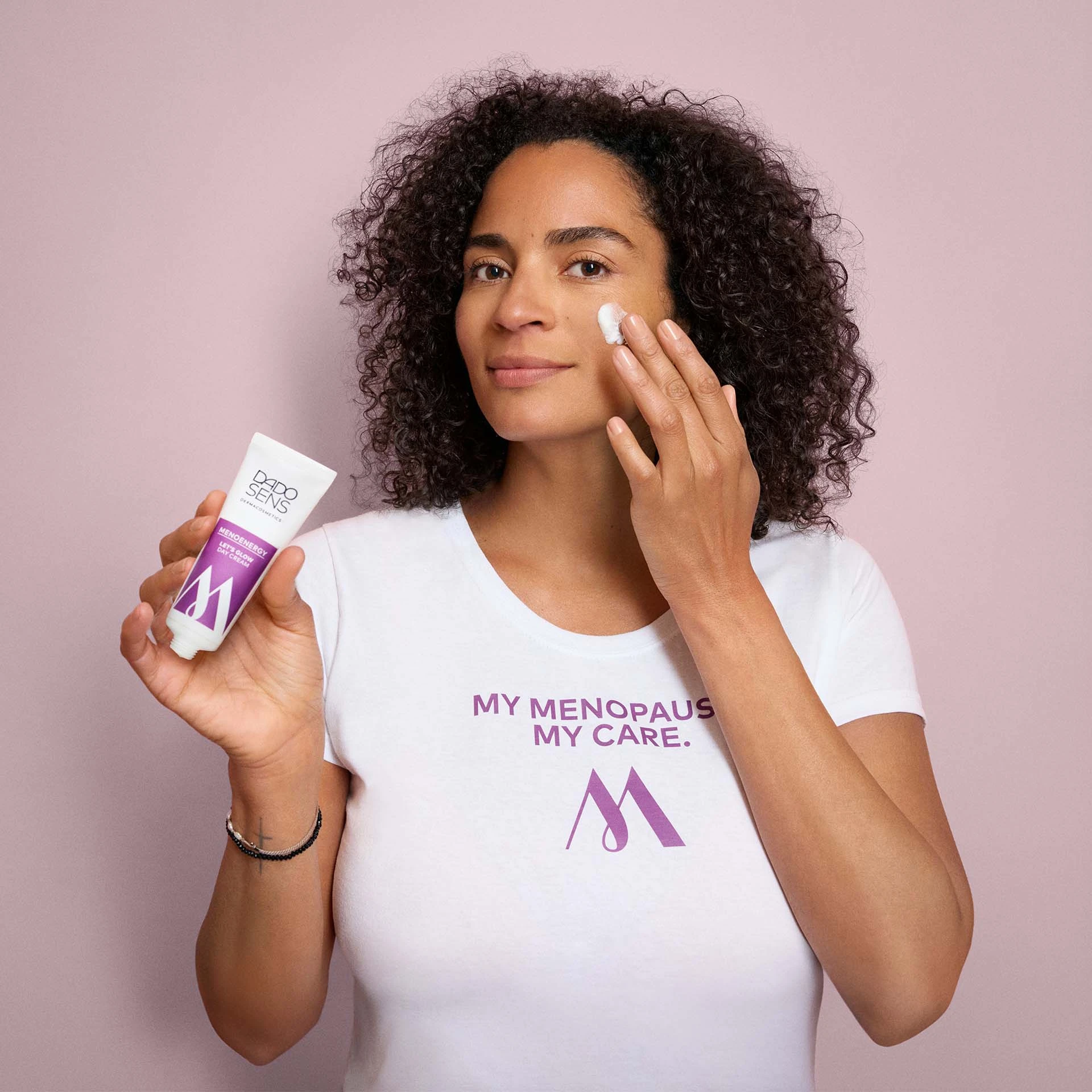 Frau trägt DADO SENS MENOENERGY LET'S GLOW Tagescreme auf Wange auf, weißes Shirt mit Slogan