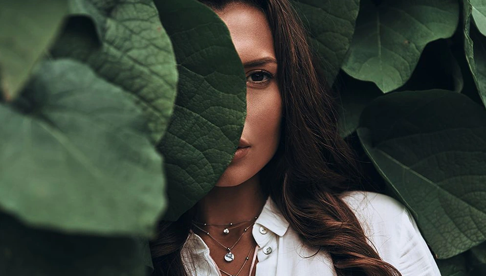 Was ist Naturkosmetik: Frau mit natürlichem Look blickt hinter großen grünen Blättern hervor – Symbolbild für die Verbindung von Schönheit und Natur.