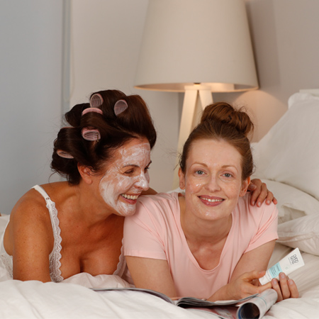 Zwei Frauen liegen im Bett, eine mit Gesichtsmaske und Lockenwicklern, die andere hält eine DADO SENS Creme