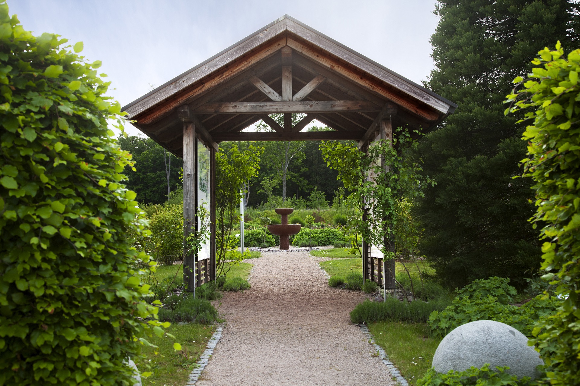 Holzpergola mit Weg, umgeben von grünen Büschen, im Hintergrund ein zweistufiger Brunnen und Wald