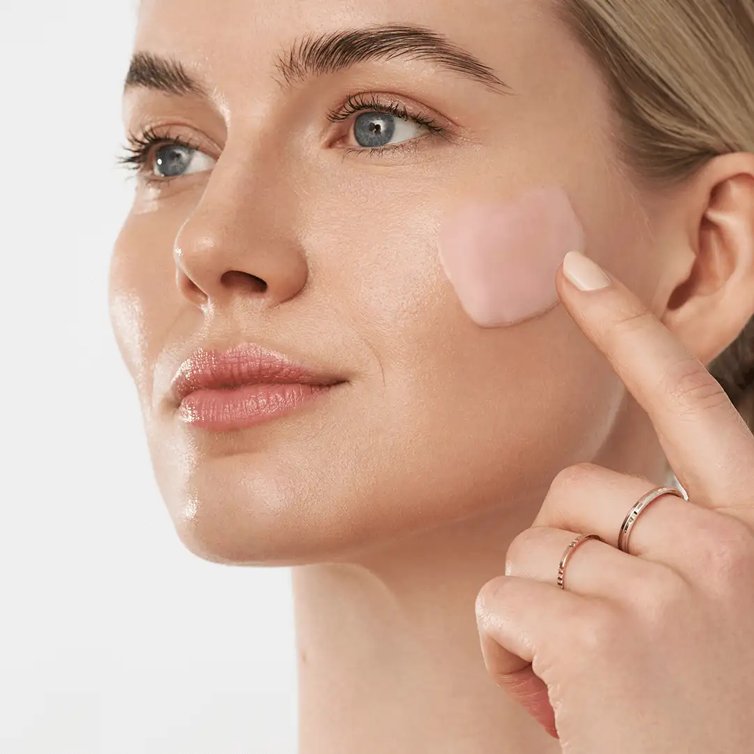 Frau trägt rosa Creme mit Finger auf Wange auf, Nahaufnahme glatter Haut und blauer Augen