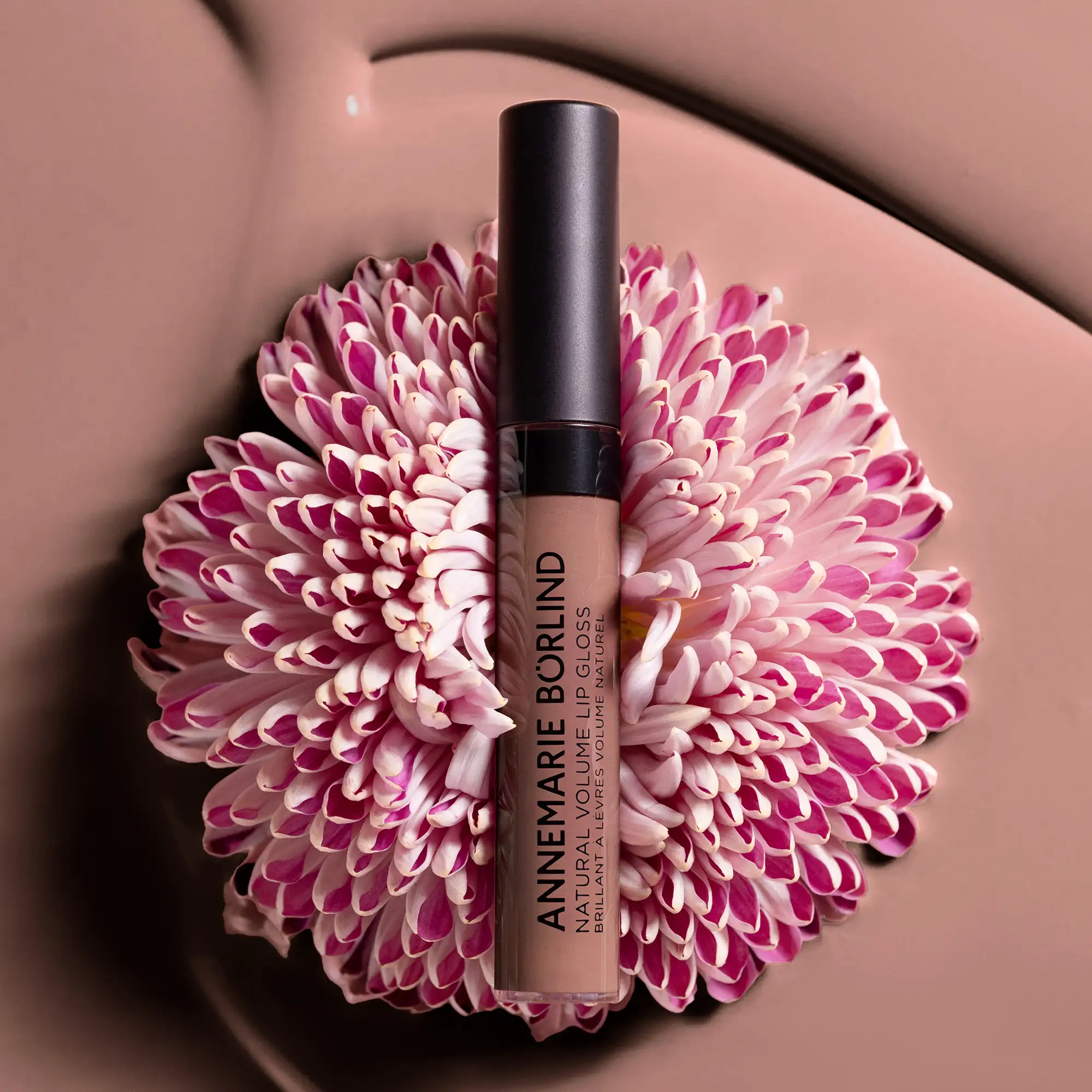 ANNEMARIE BÖRLIND NATURAL VOLUME LIP GLOSS in Farbe Nude Hug, dekorativ auf Blüte mit rosa und weißen Blütenblättern vor braunem Hintergrund inszeniert