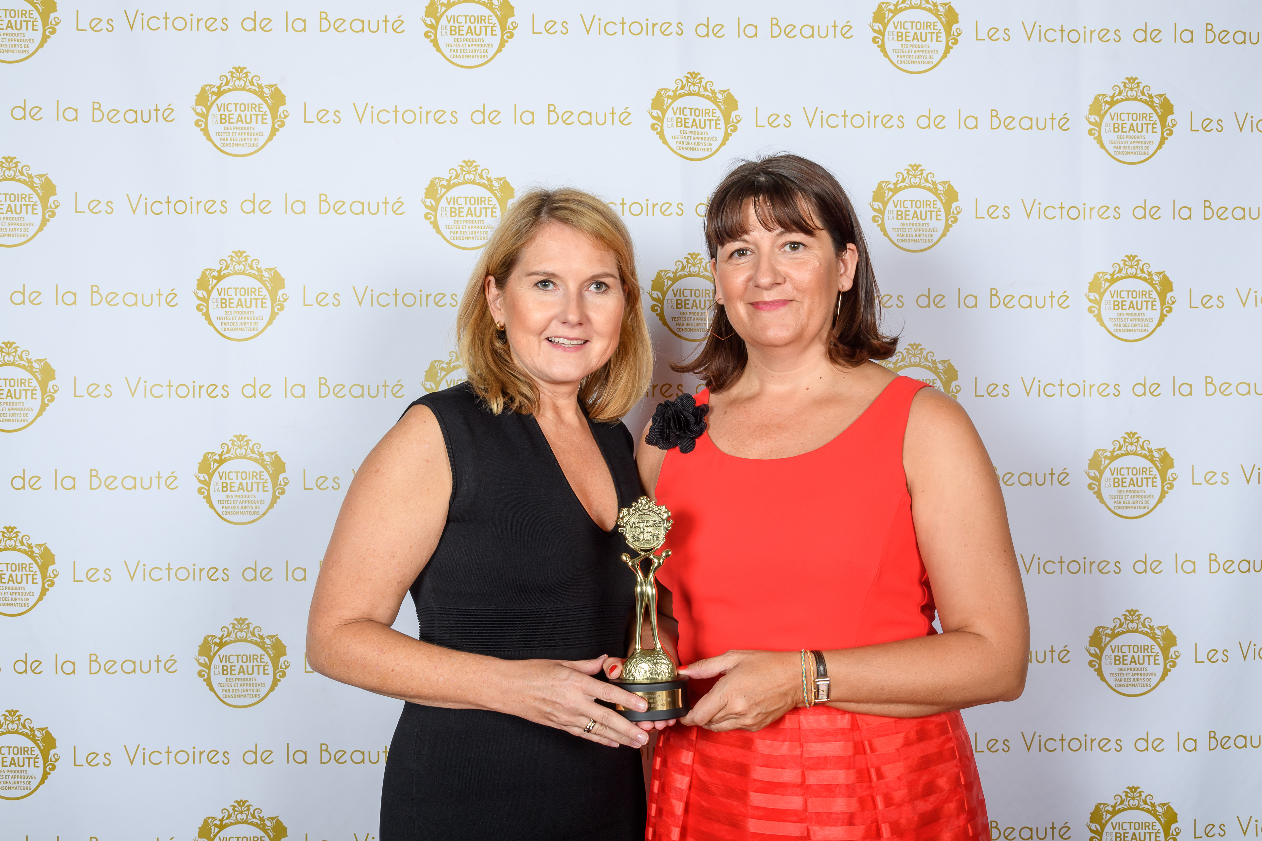 Zwei Frauen halten gemeinsam eine goldene Trophäe vor einem Hintergrund mit "Les Victoires de la Beauté"-Schriftzug