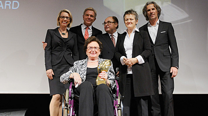 Sechs Personen in Businesskleidung, eine Frau im Rollstuhl hält eine goldene Trophäe bei Preisverleihung