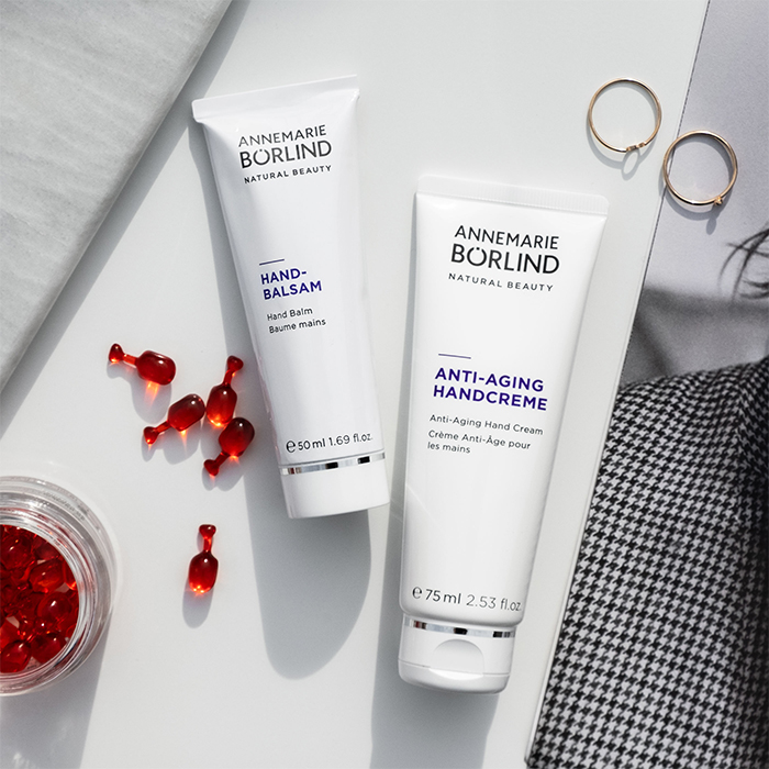 ANNEMARIE BÖRLIND Handbalsam 50 ml und Anti-Aging Handcreme 75 ml auf weißem Untergrund mit Schmuckringen und roten Kapseln