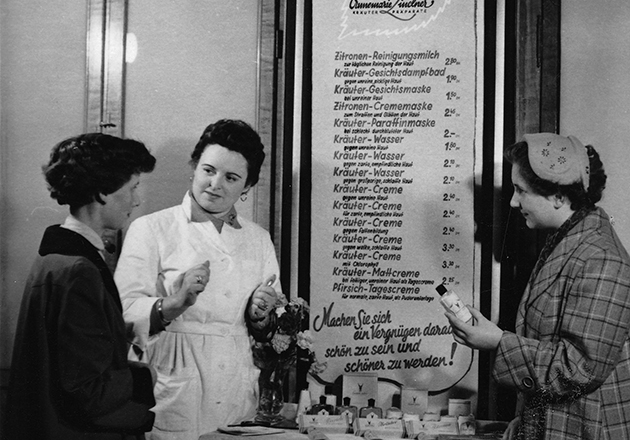Annemarie Lindner und zwei Frauen vor Preisliste für kosmetische Behandlungen und Produkte, eine berät die anderen, 1950er Jahre.
