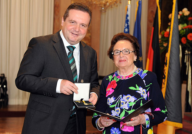 Zwei Personen bei feierlicher Auszeichnung, Mann hält Medaille, Frau hält Urkunde, Flaggen im Hintergrund