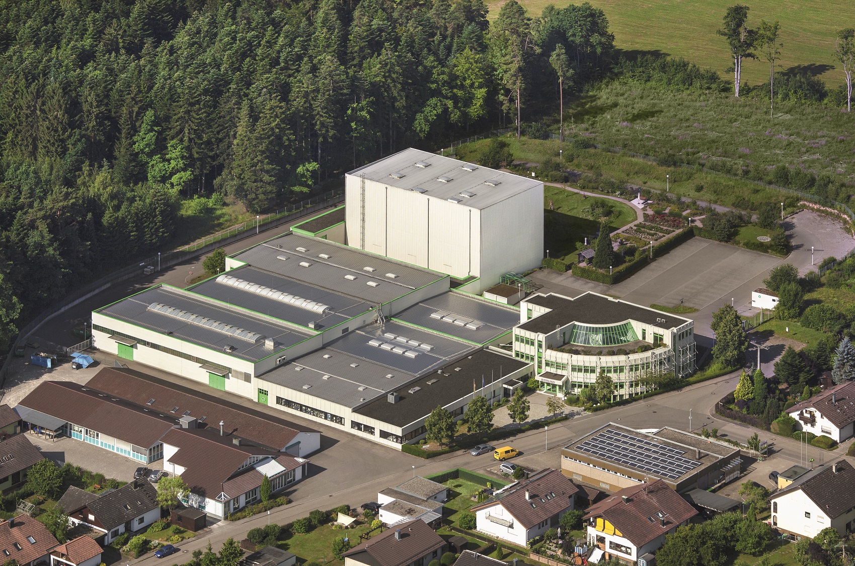 Luftaufnahme eines Industriegebäudes mit Bürotrakt, umgeben von Wohnhäusern und Wald im Hintergrund